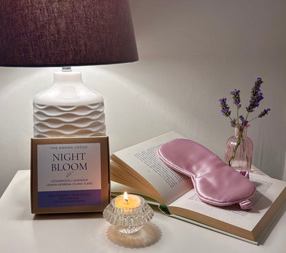 Pink eye mask on an open book with a candle, lamp, and lavender flowers in the background.