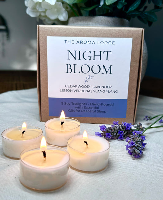 Three lit candles in white holders with a box labeled 'The Aroma Lodge Night Bloom' on a light surface.