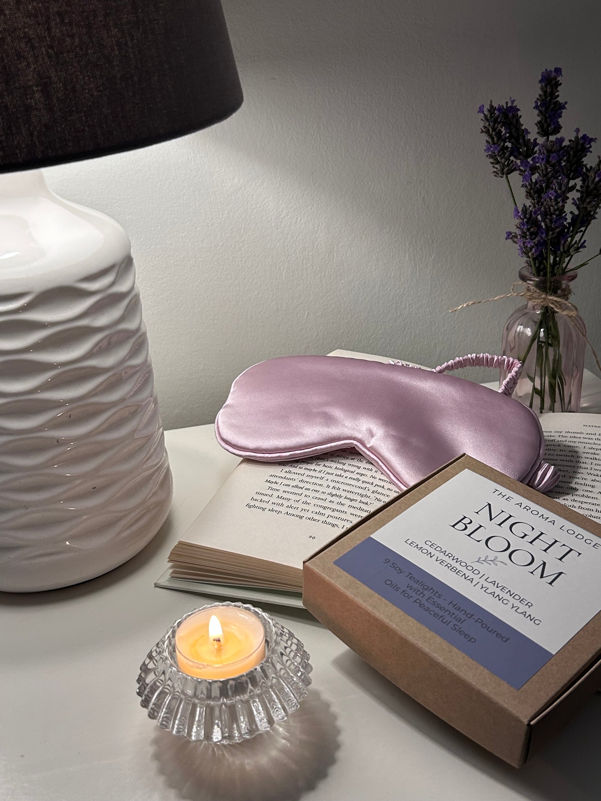 Pink satin eye mask on a book with a candle and lavender flowers on a gray surface